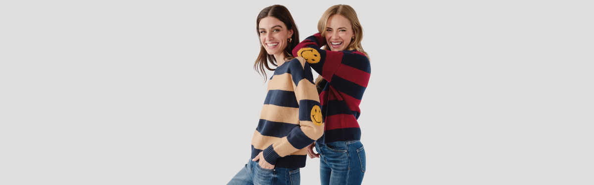 two women smiling wearing striped sweaters and jeans with smiley faces on the elbows, leaning on each other