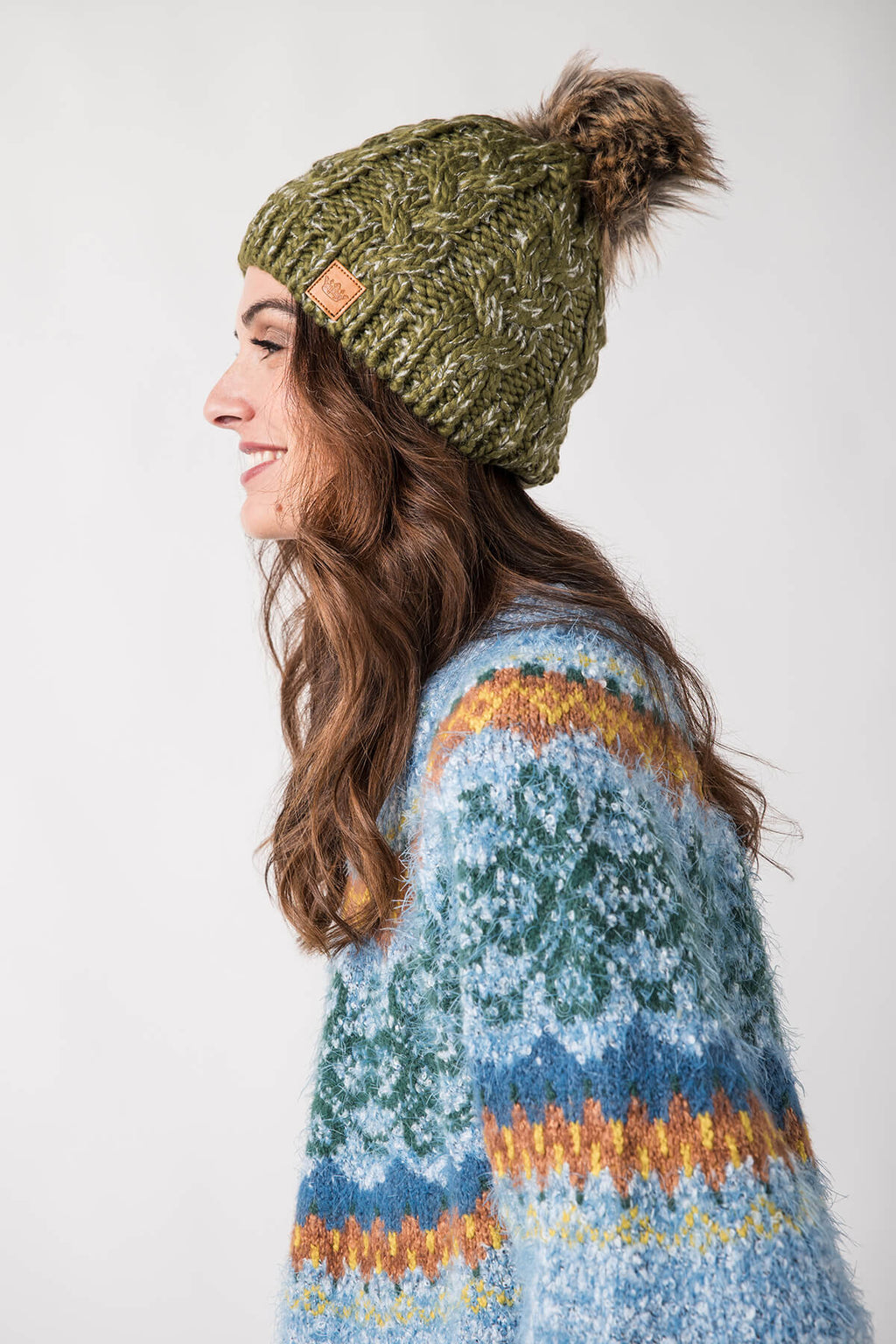 Olive cable-knit pom hat with ribbed cuff, side knit patch, and fluffy pom.